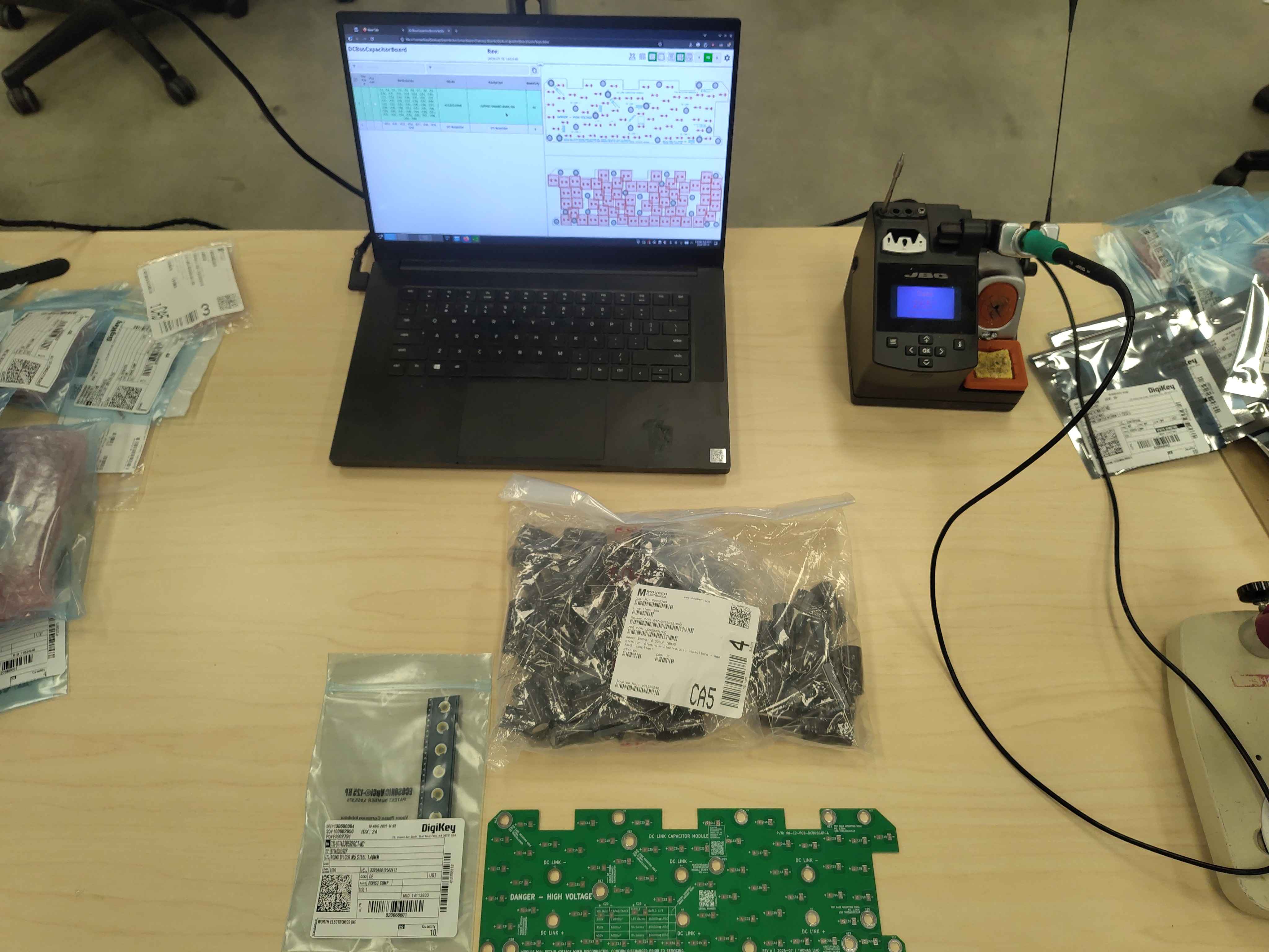 Workspace with PCB, capacitor reel, standoffs, soldering station, and BOM on a laptop